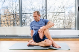 Sur un tapis posé dans son studio, un homme portant des vêtements de yoga effectue une posture complexe.