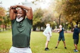 Muscular man stretching his arms while others stretch in the background at a public park.
